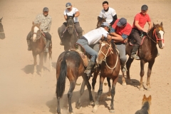 Two riders compete with one another to try and wrestle each other off the horse snatching up a dead goat from the ground while the horses gallop at top speed.