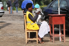 A young mother and her baby sitting near a street.   She has a yellow vest on and that signifies what she is selling.