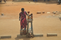 A young girl selling bootleg Gasoline using glass bottles to pour it in by hand into the gasoline tanks.  For motorbikes mainly.